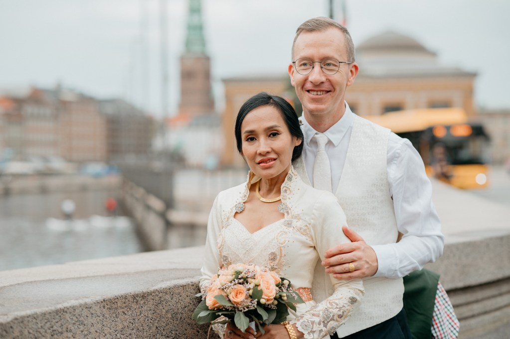 A bride and groom stand together on a stone bridge in an urban setting. The bride wears a traditional off-white outfit with intricate embroidery and holds a bouquet of peach roses with greenery. She is accessorized with gold jewelry. The groom, dressed in a white shirt, tie, and embroidered waistcoat, smiles while holding the bride gently. The background features blurred city architecture, a canal, and a yellow bus, creating a romantic and serene atmosphere.