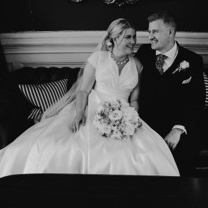 A bride and groom sitting closely together on a vintage sofa, smiling warmly at each other on their wedding day. The bride wears a classic satin gown and holds a floral bouquet, while the groom is dressed in a formal suit.