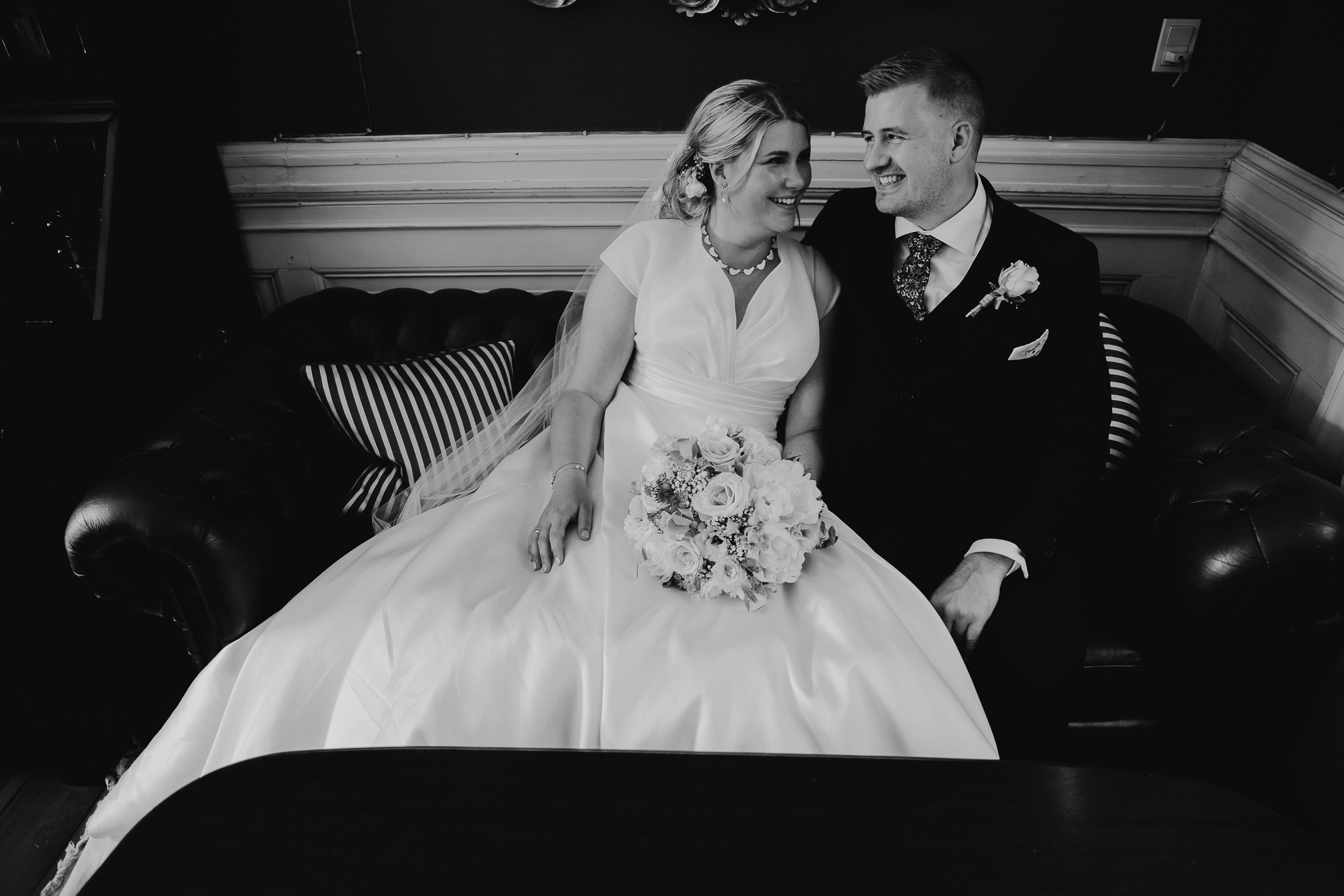 A bride and groom sitting closely together on a vintage sofa, smiling warmly at each other on their wedding day. The bride wears a classic satin gown and holds a floral bouquet, while the groom is dressed in a formal suit.