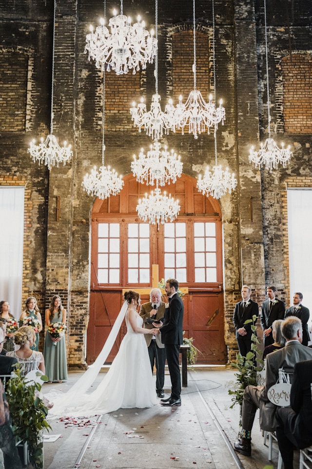 A bride and groom exchange vows in a stunning indoor wedding venue with rustic brick walls and large wooden doors in Denmark. The space is illuminated by elegant chandeliers, creating a warm and romantic atmosphere. The bridal party and guests look on as the couple shares this beautiful moment, perfect for a memorable wedding celebration in Denmark.