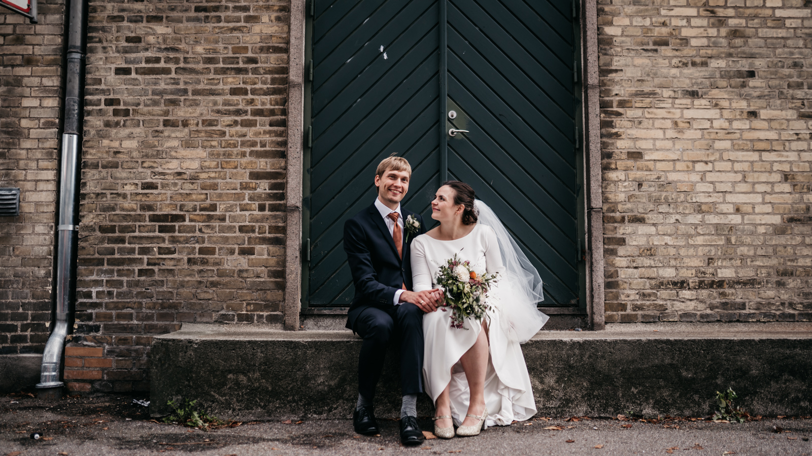 A bride and groom sit together on a stone ledge in front of a rustic brick wall with a dark green door. The bride wears a long-sleeved white dress and veil, holding a bouquet, while the groom is dressed in a dark suit with a warm-colored tie. They are smiling warmly at each other, creating an intimate and joyful moment. Perfect for capturing the charm of a Danish wedding setting.