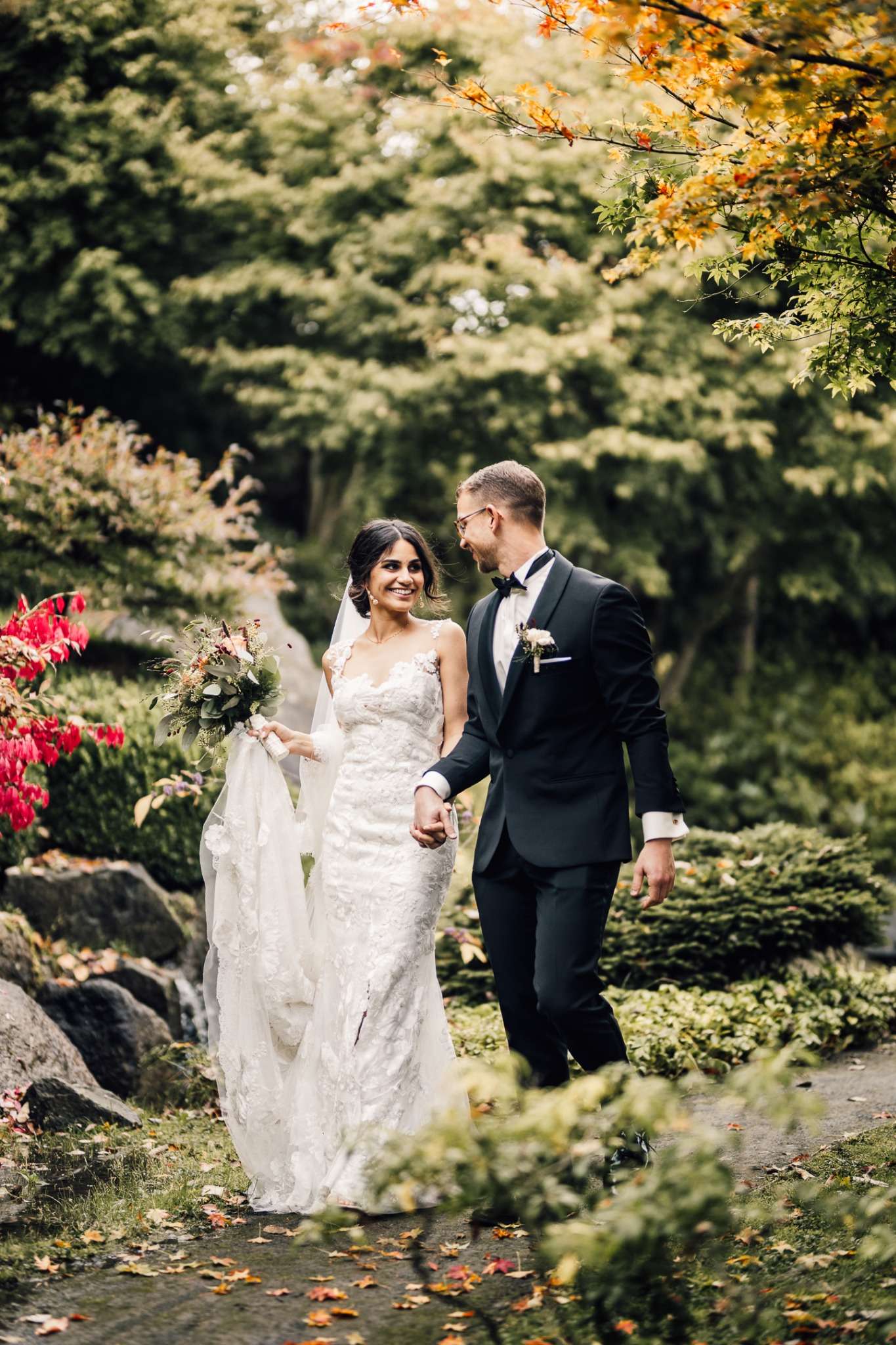 A bride and groom walk hand in hand through a beautiful, lush garden in Denmark. The bride wears a lace wedding dress and holds a bouquet of greenery and flowers, while the groom is dressed in a classic black tuxedo. They smile warmly at each other, surrounded by vibrant autumn foliage, capturing a romantic and intimate moment in a scenic natural setting. Perfect for a wedding in Denmark.