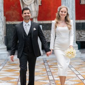 A bride in a white dress and groom in a black suit standing in a decorated hall with marble floors and red walls.