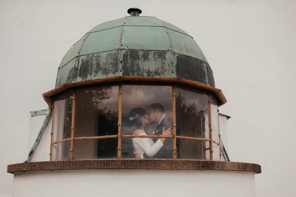 Captured through antique glass, Greg and Anghela kiss inside the lighthouse’s old domed room, surrounded by rustic charm and timeless romance