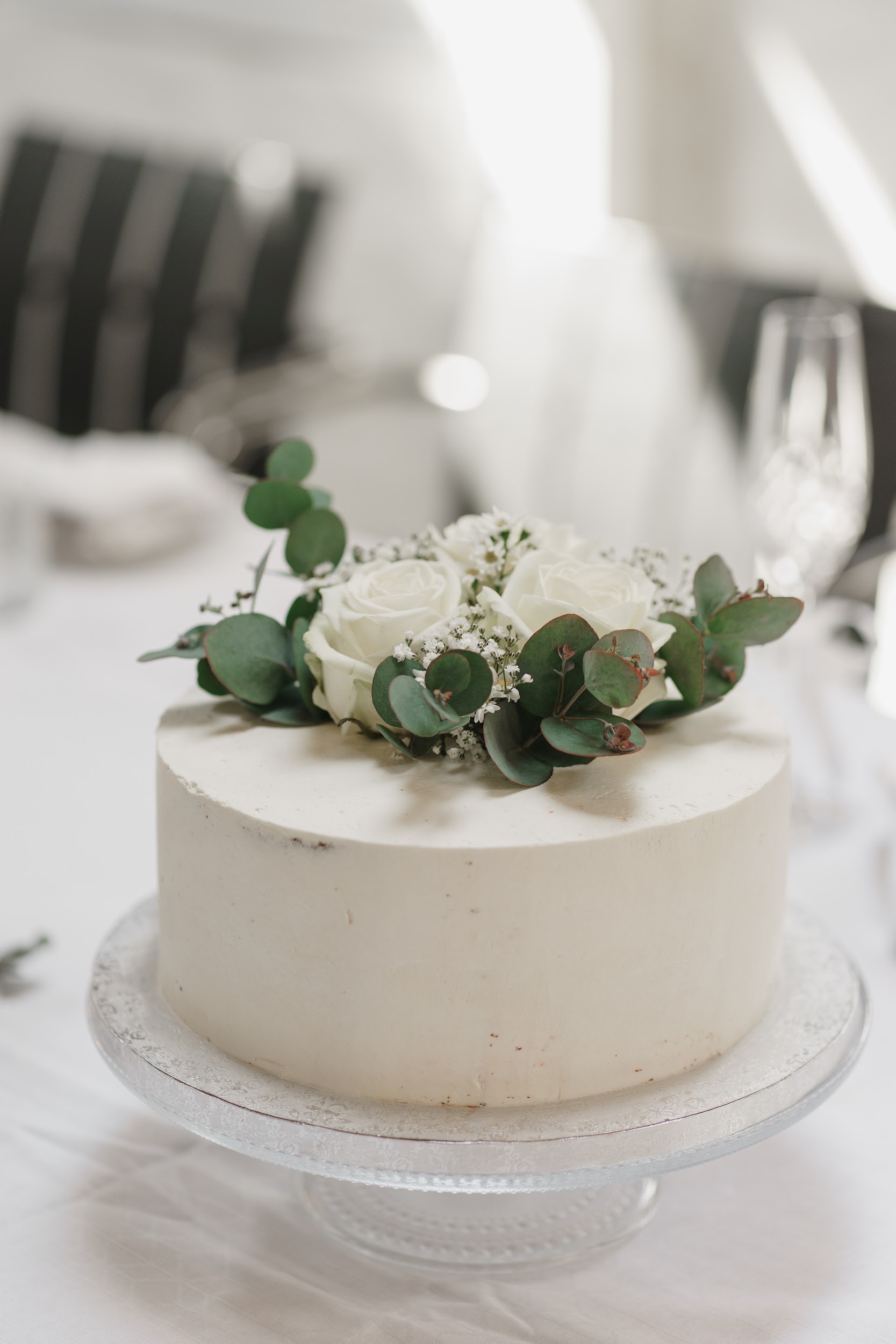 Simple white wedding cake decorated with white roses and eucalyptus on a glass cake stand.