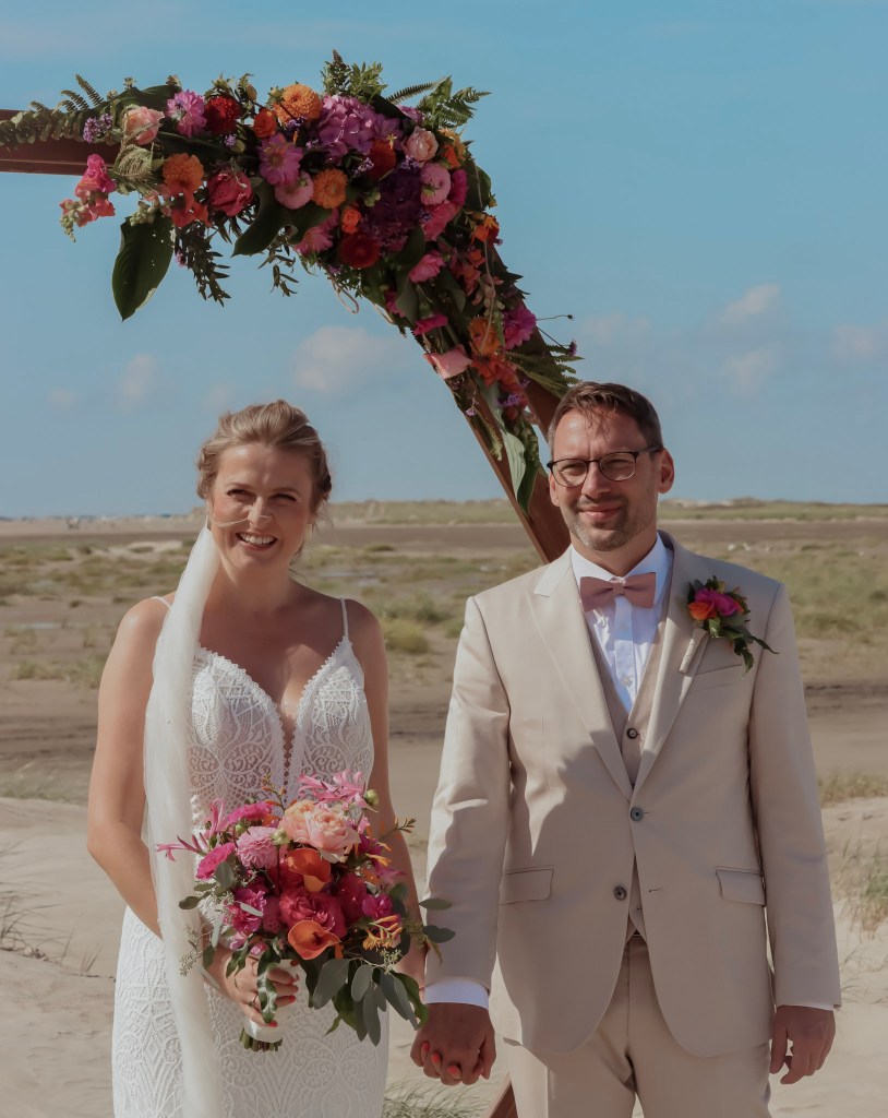 Bride and groom standing under a floral arch on the beach.