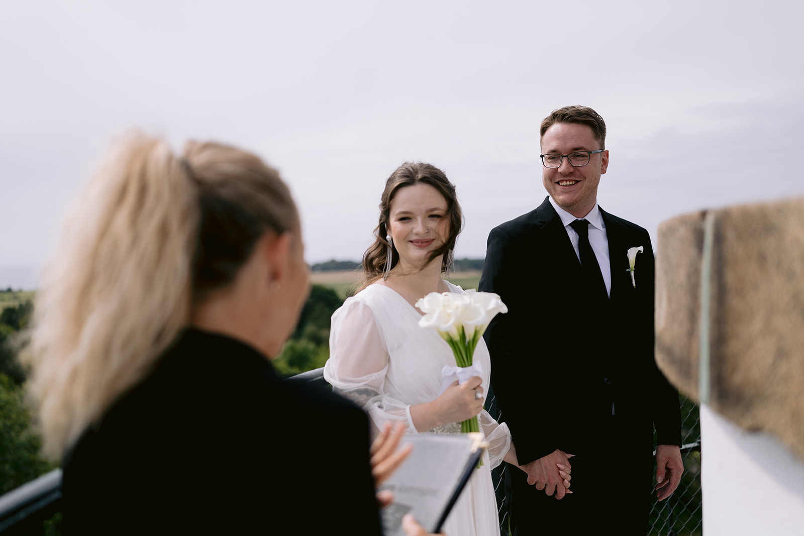 A bride and groom holding hands during their outdoor wedding ceremony, with the bride holding a bouquet of white flowers and smiling at the officiant.