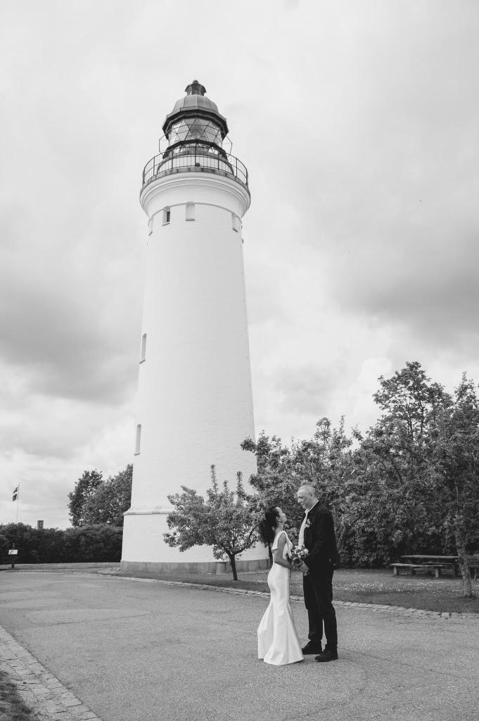 Bride and groom standing in front of a tall white lighthouse, smiling at each other.