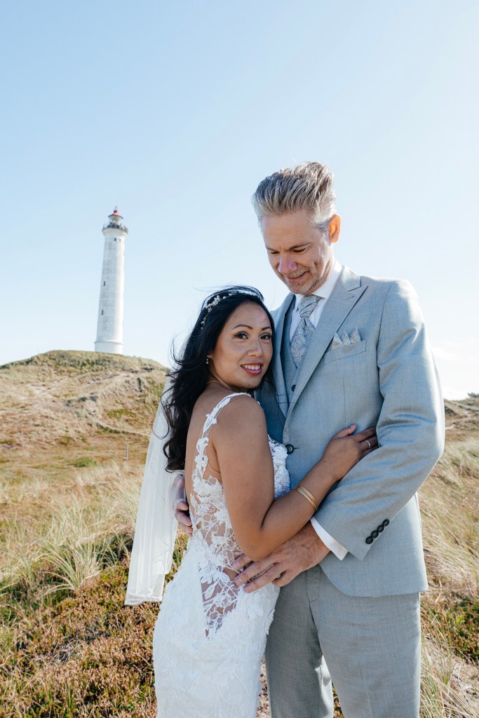 Bride and groom embracing in front of a lighthouse on a sunny day.