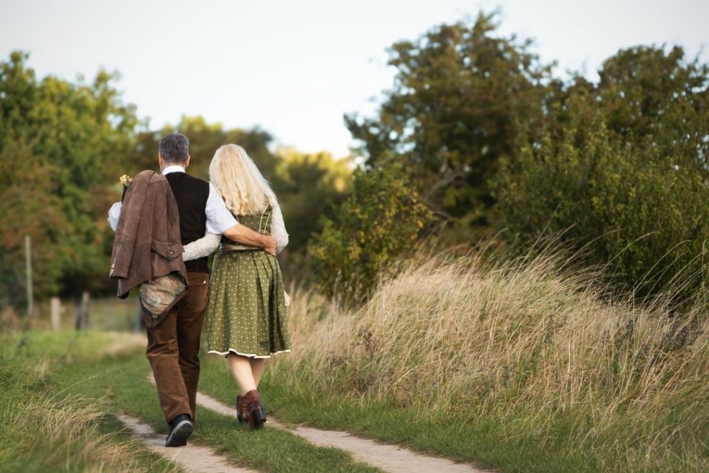A couple walks arm in arm down a rural path surrounded by tall grass and trees.