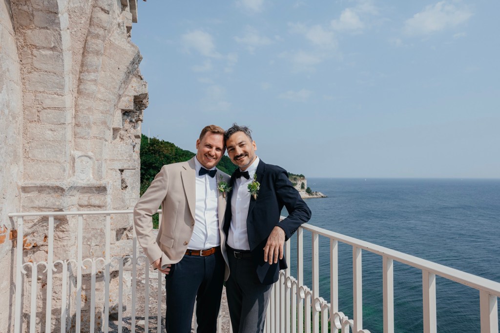 Same-sex couple smiling after their wedding in Denmark at a scenic coastal location with sea views and historic architecture.